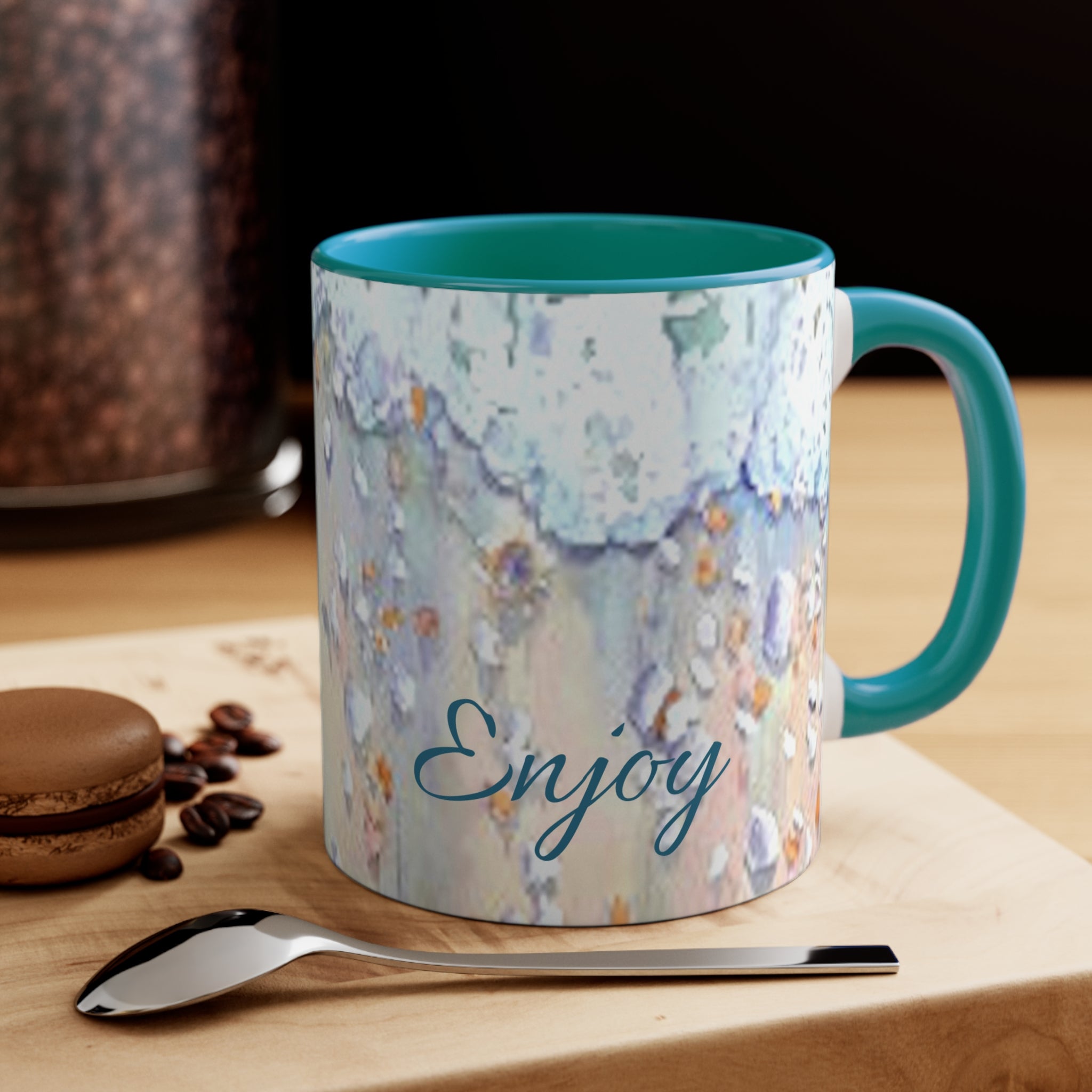 Mug with floral design and 'Enjoy' text on a wooden surface with coffee beans and a spoon.