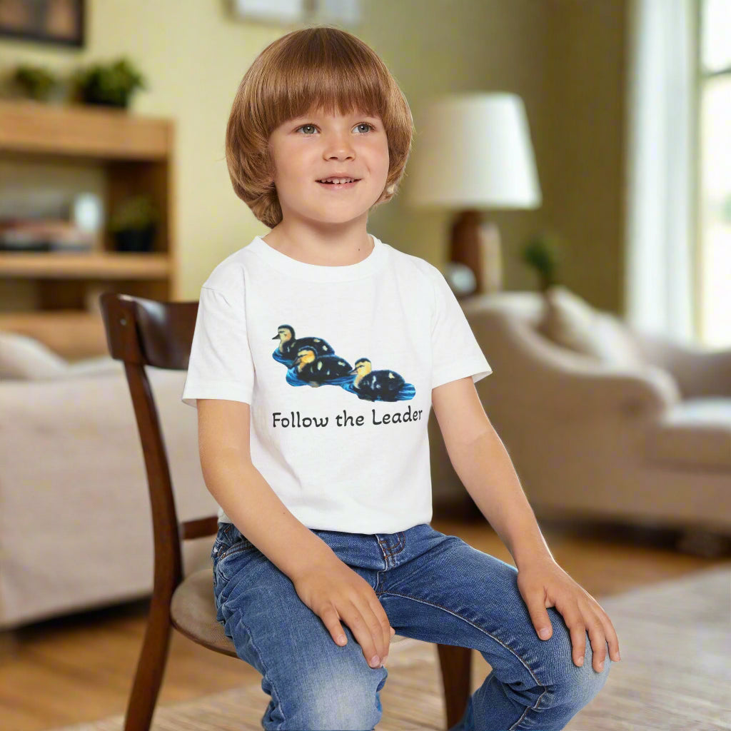 Child wearing a white t-shirt with a graphic and text, sitting on a white stool against a light wooden background.
