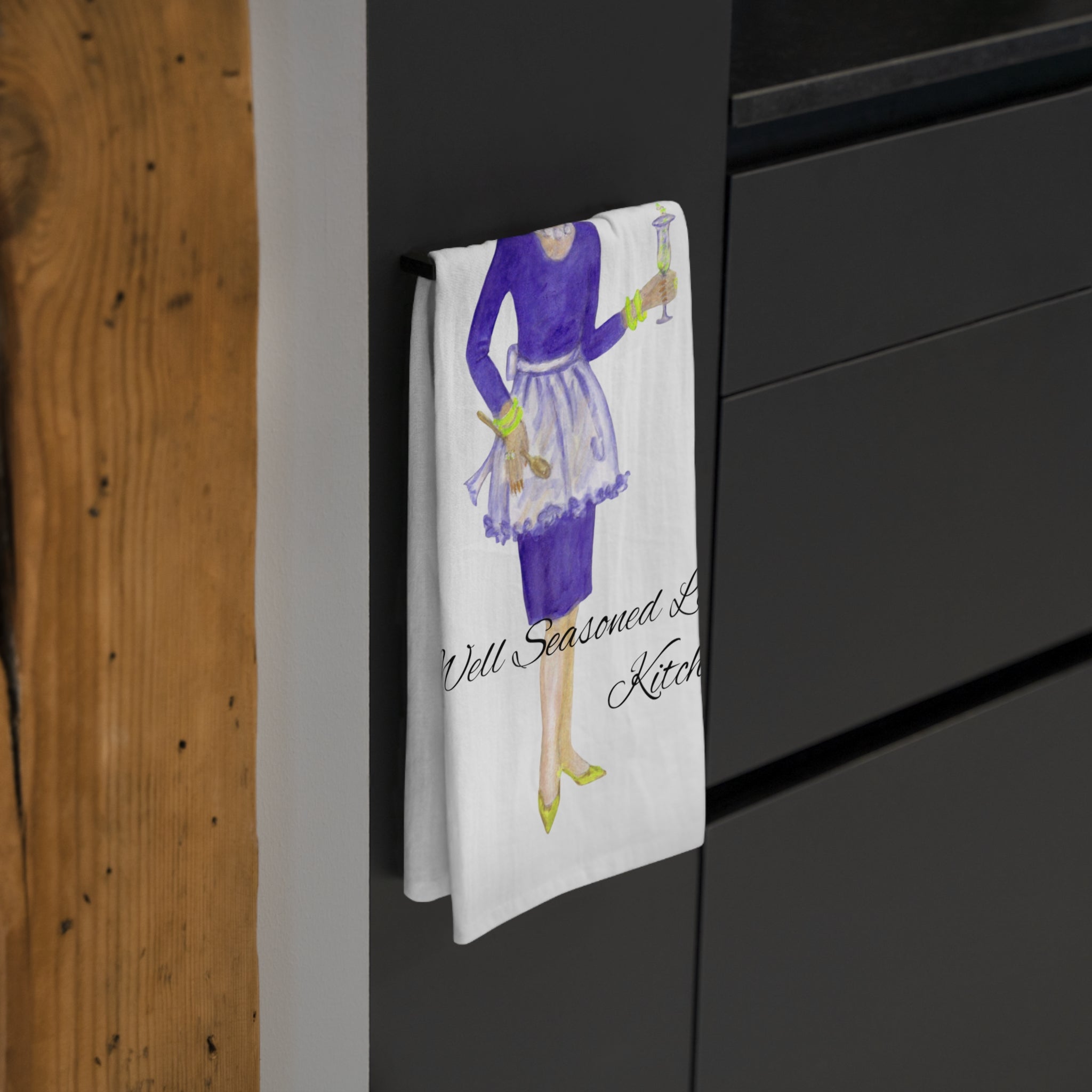 Tea towel with a design of a woman in a kitchen hanging on a black cabinet.
