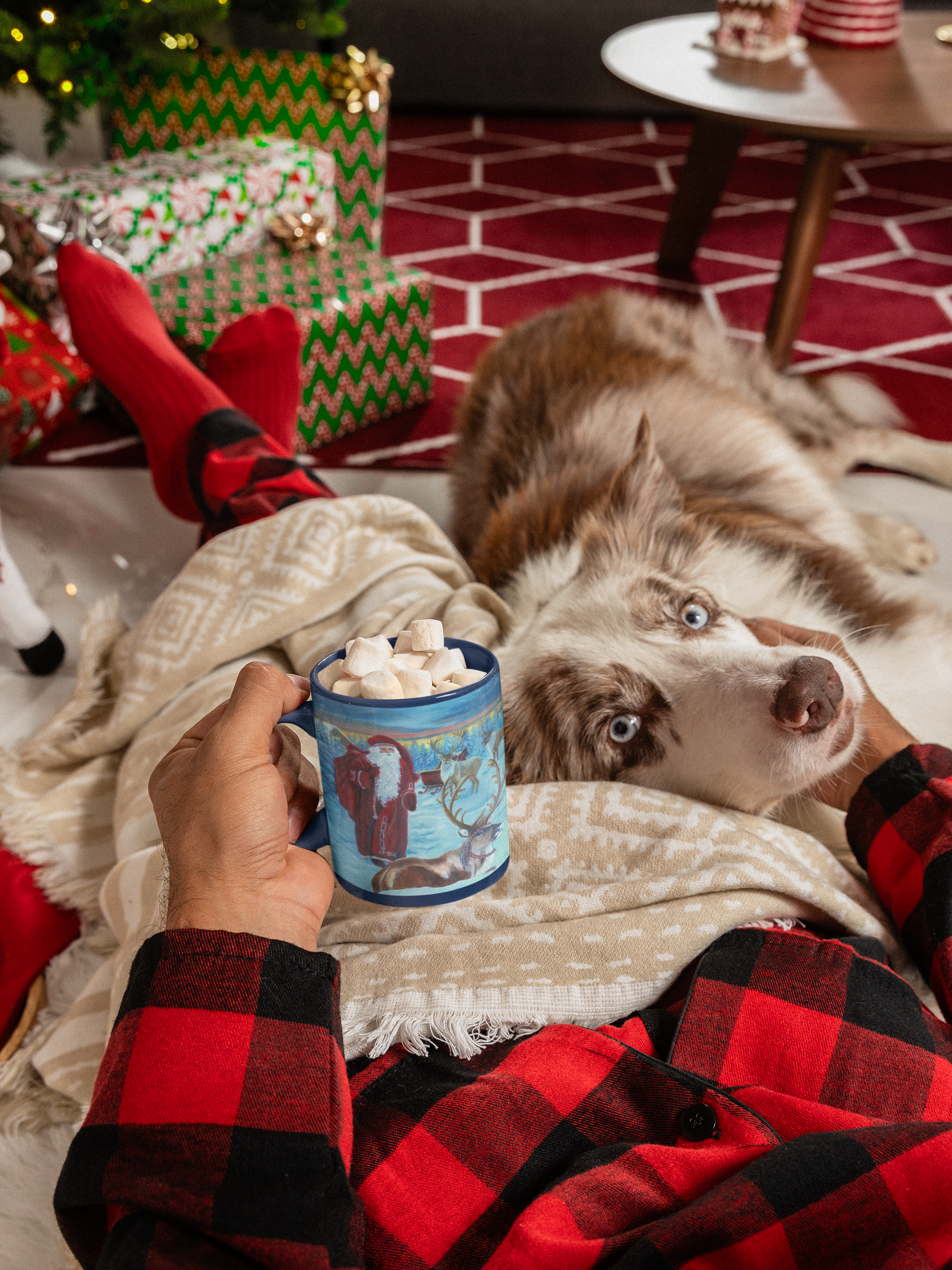 Person in red plaid pajamas holding a mug with a dog lying next to them on a cozy rug.