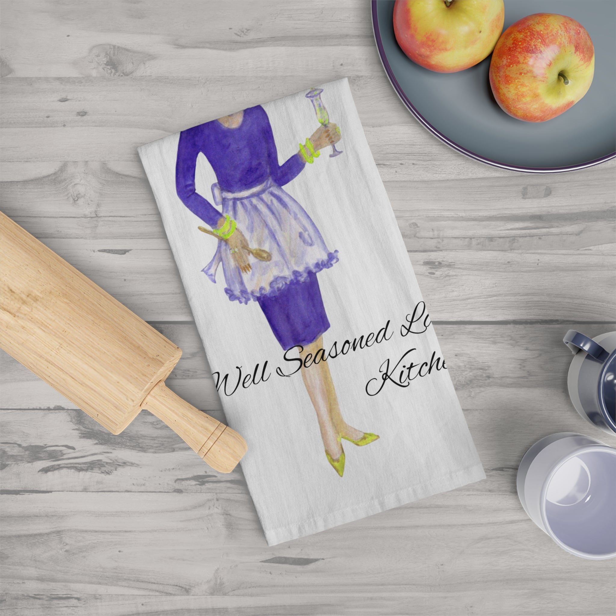 Kitchen towel with a design of a woman in a kitchen, placed on a wooden surface with apples and a rolling pin.