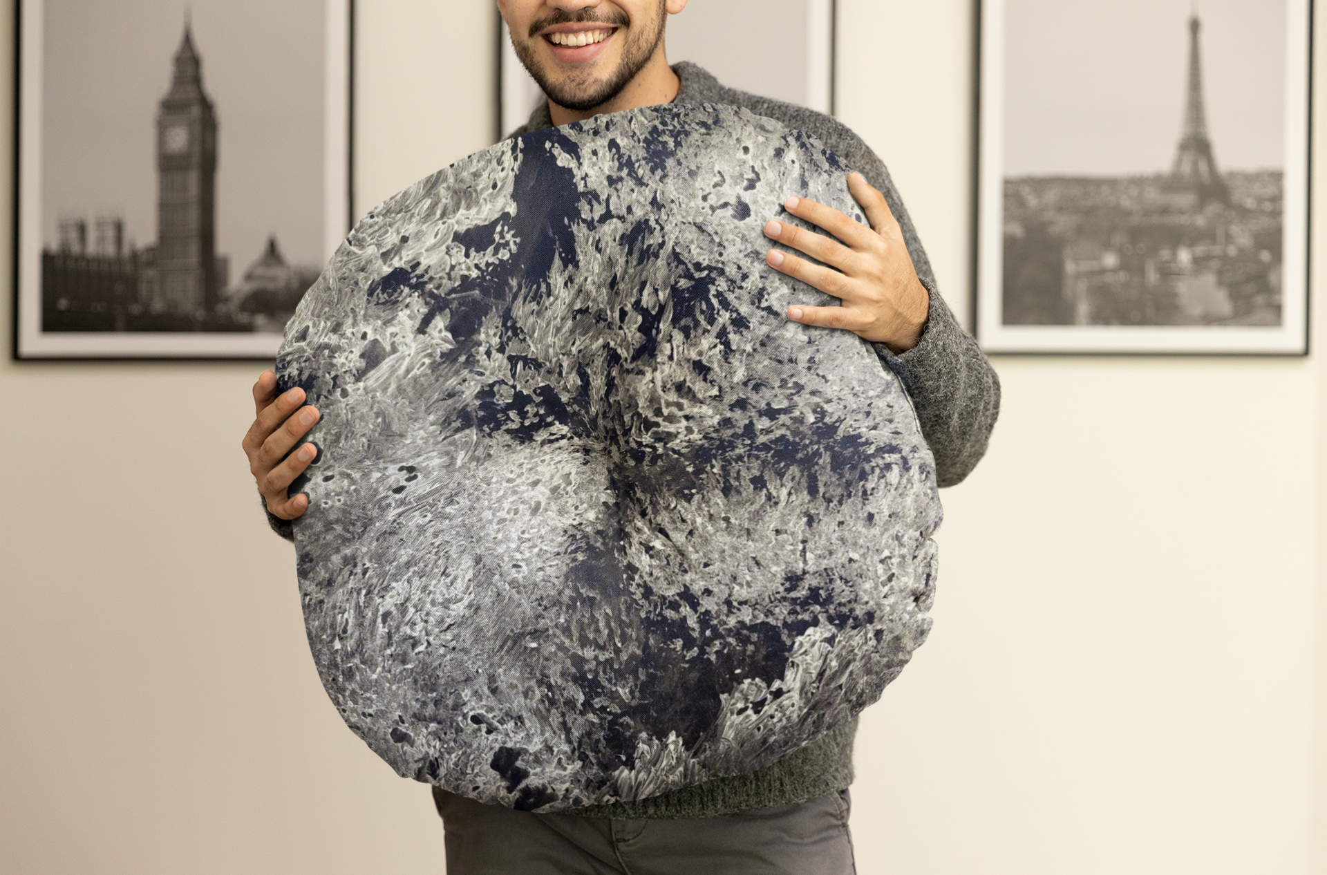 Person holding a large moon-shaped pillow in a room with framed pictures on the wall.
