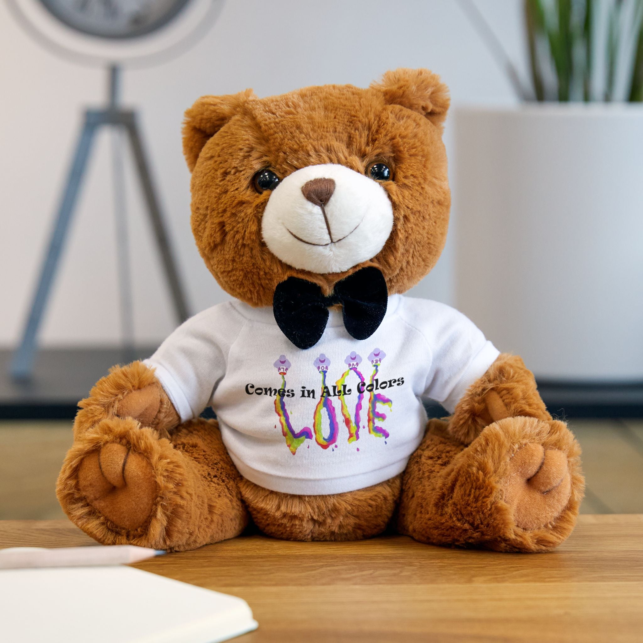 Brown teddy bear wearing a white shirt with colorful text on a wooden surface.